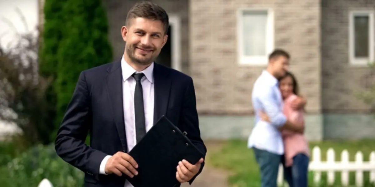Conseiller en costume tenant un porte-bloc devant une maison, un couple enlacé à l'arrière-plan